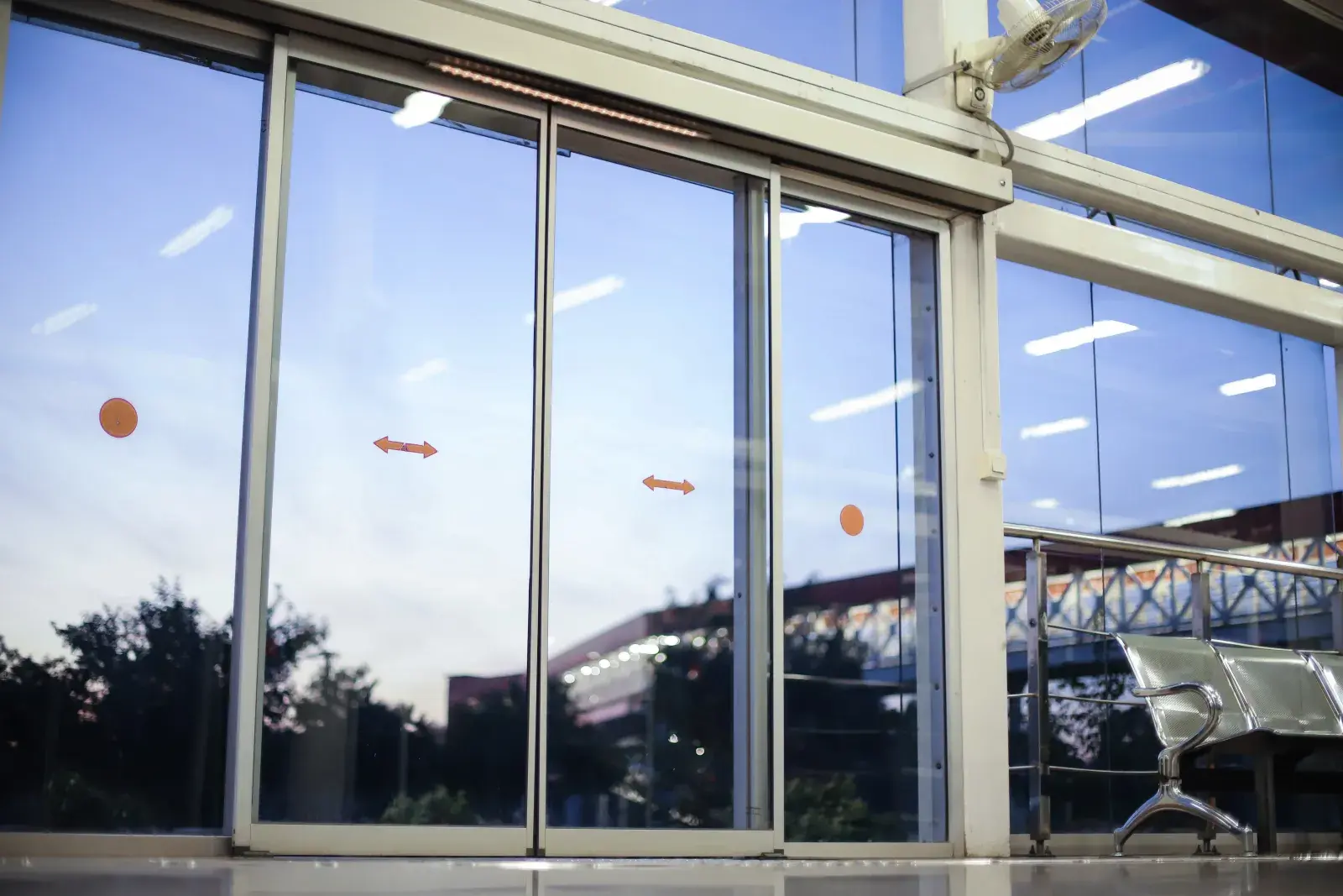 Puertas automáticas de cristal en una estación de transporte, con señalización naranja y bancos de espera metálicos a un lado.
