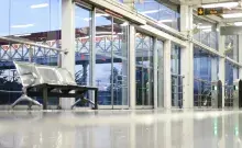 Vista a nivel de suelo de una estación de transporte, mostrando el reflejo del interior, bancos metálicos y puertas automáticas de cristal.