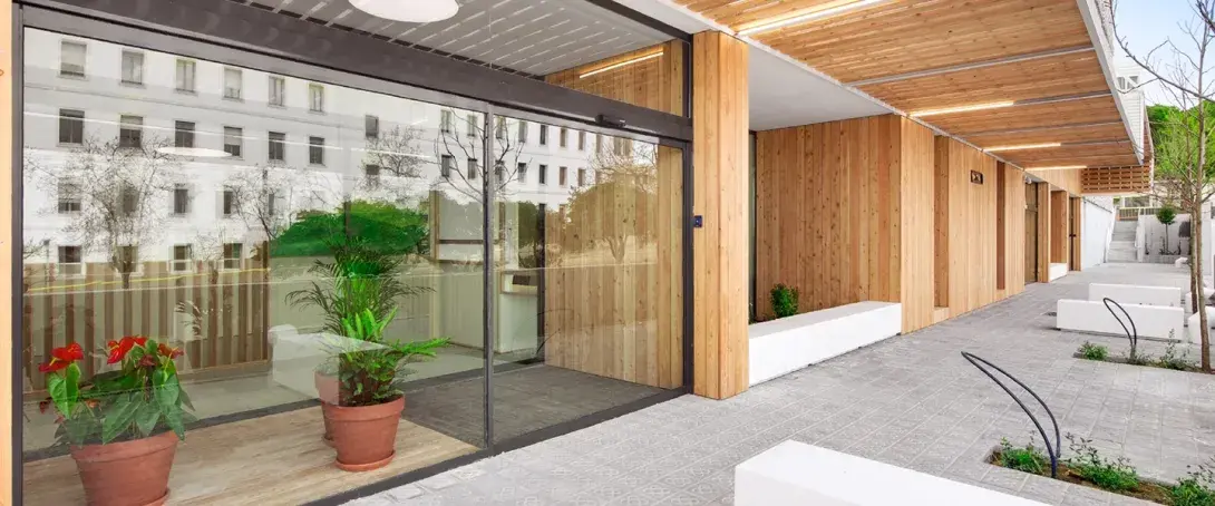 Puerta automática corredera de cristal en la entrada de un edificio moderno con fachada de madera y acceso exterior ajardinado.